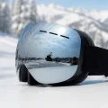 Silver mirror snow goggles shown at an angle on snow with a black head strap