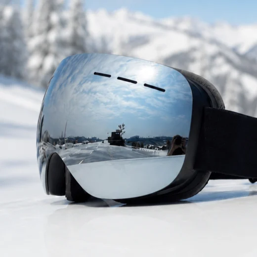 Silver mirror snow goggles shown at an angle on snow with a black head strap